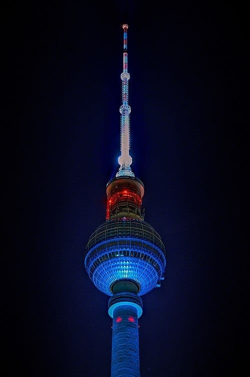 The top of the Berlin Television Tower.  It is a thin metallic structure ending in a sphere and spire with red lights.  There is black sky behind it.