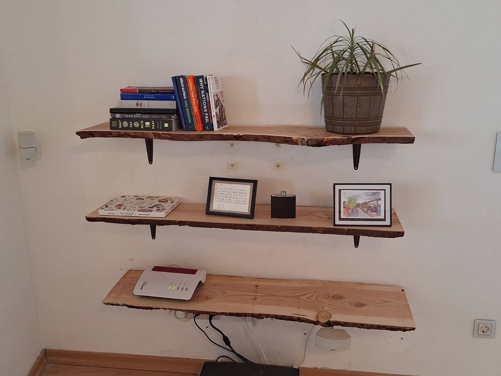 Three wooden wall-mounted shelves made from natural rustic-looking wood planks.  Top shelf contains a group of books on the left, including "Why Nations Fail" and "Salt Fat Acid Heat", and a potted plant with long, narrow leaves in a wooden-style pot on the right.  Middle shelf has framed cards, one is just text and one is a street scene, and a black whisky flask - the text and flask were presents from my best friend when I was best man at his wedding. Bottom shelf has a wifi router. The wall is white, and the shelves are supported by brown metal brackets.