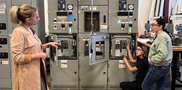 An instructor talks to two students in an Electrical Power and Controls lab setting.