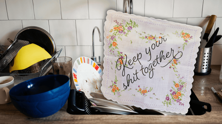 A sink full of dirty dishes and a handkerchief that says "Keep your shit together" in script font with flowers 