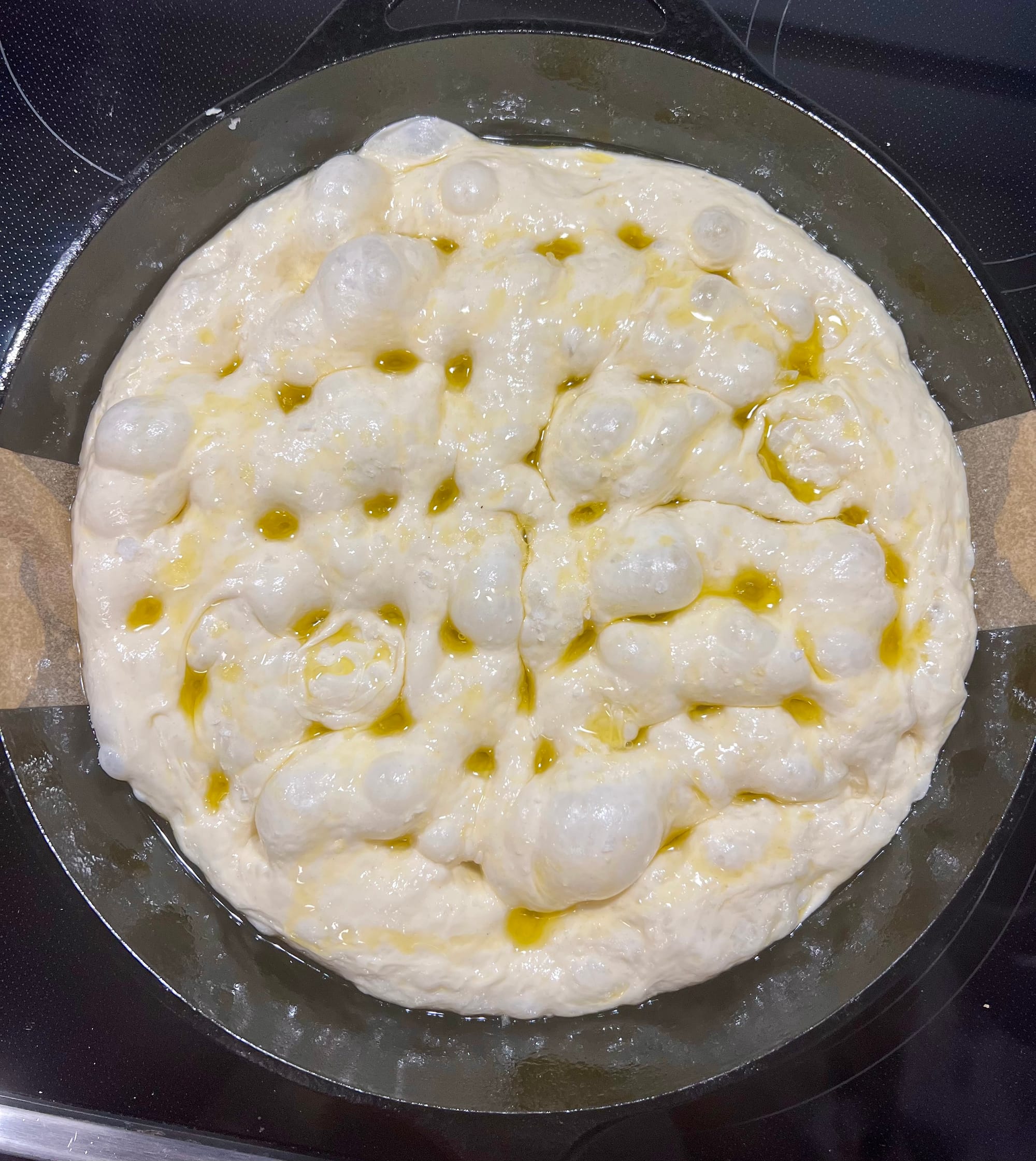 The uncooked focaccia is in a round cast-iron skillet. The surface has been dimpled by fingertips, and there is olive oil pooled on top.