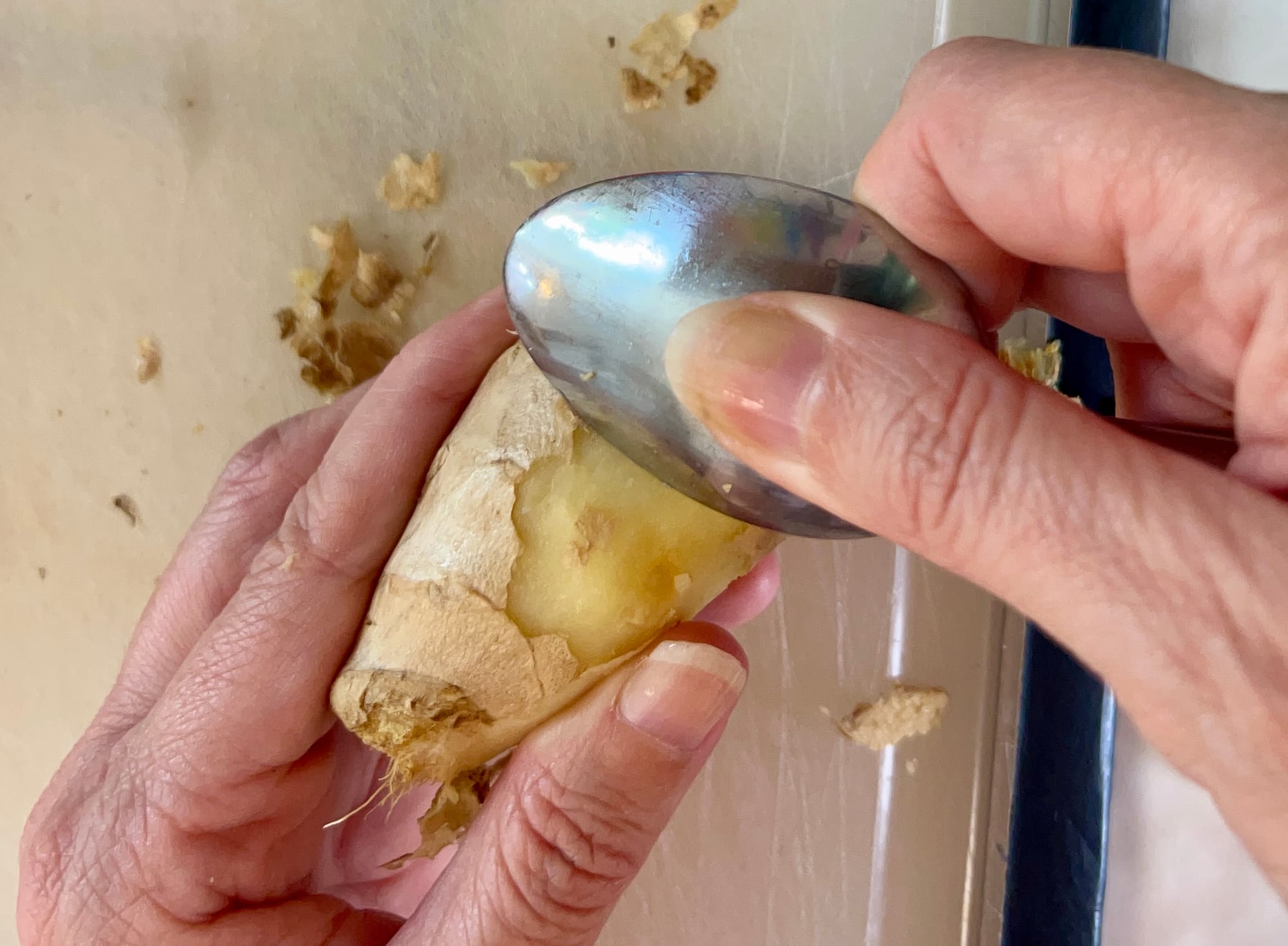 Holding a hunk of ginger with one hand, while the other scrapes the edge of a spoon along the side to remove the skin.