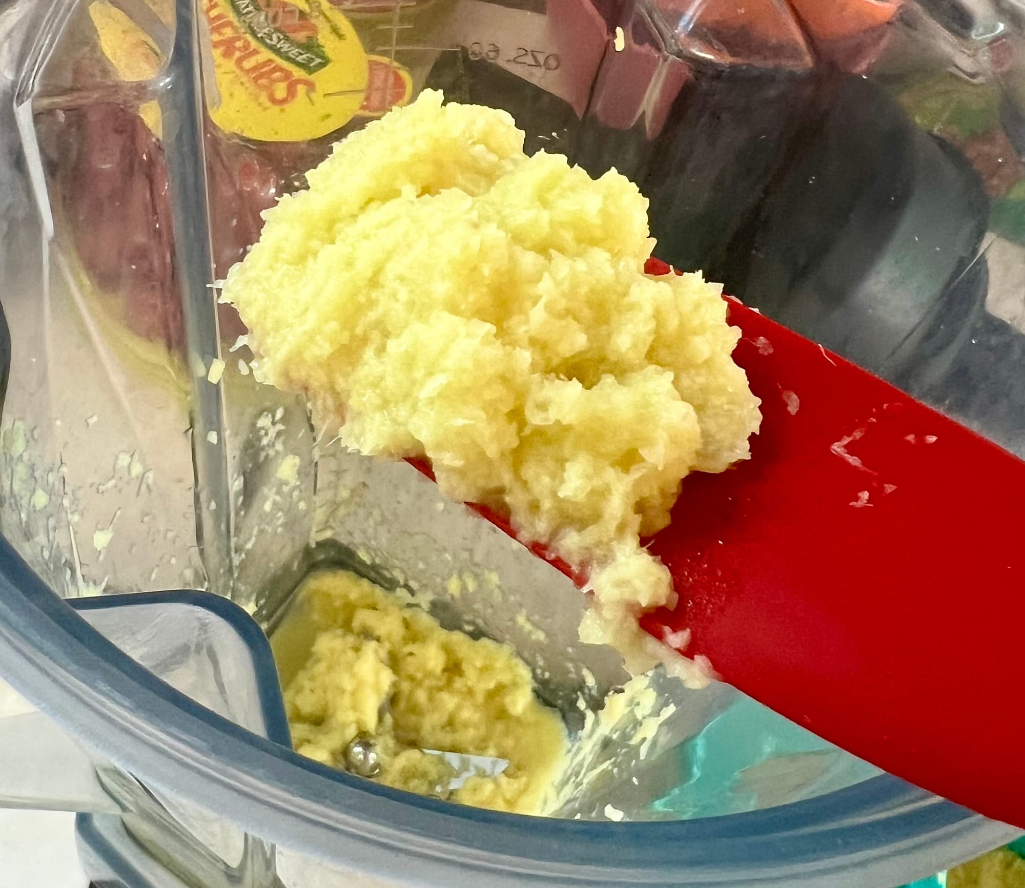 A red silicone spatula is holding up a blob of finely grated ginger from the blender.