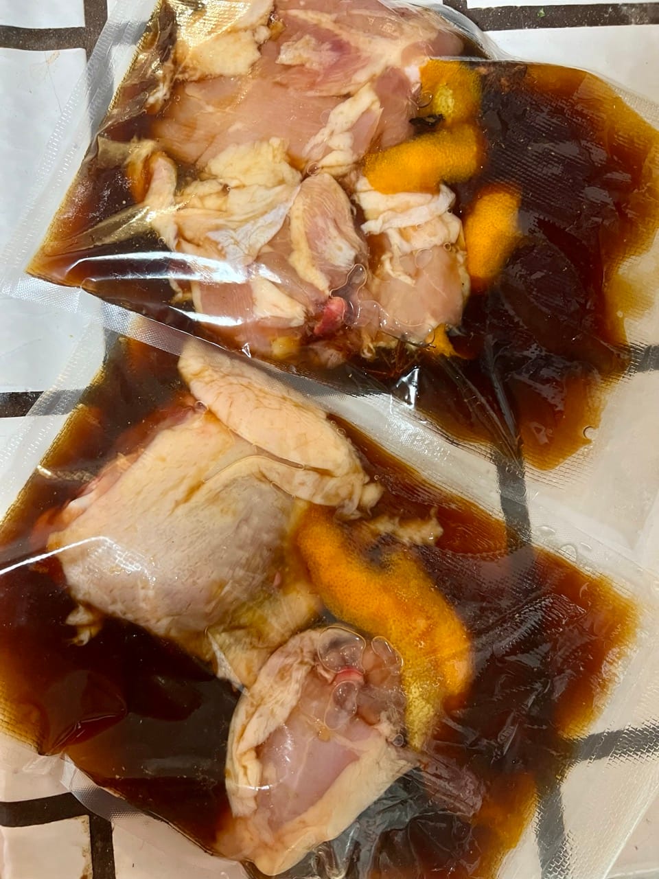 Two large plastic bags have raw chicken breasts in a dark brown marinade with orange peels.