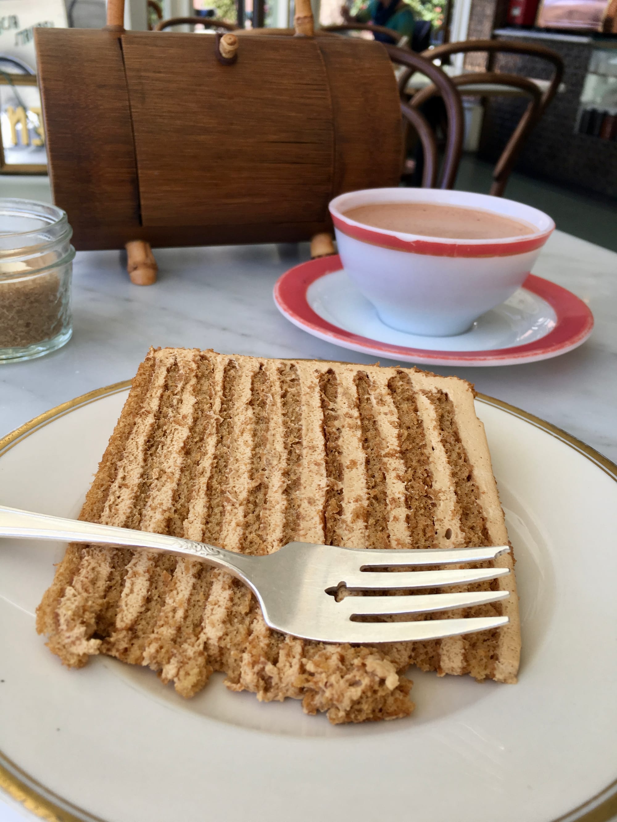 A slice of 10-layer honey cake is on a plate at a cafe, next to a teacup with hot chocolate.