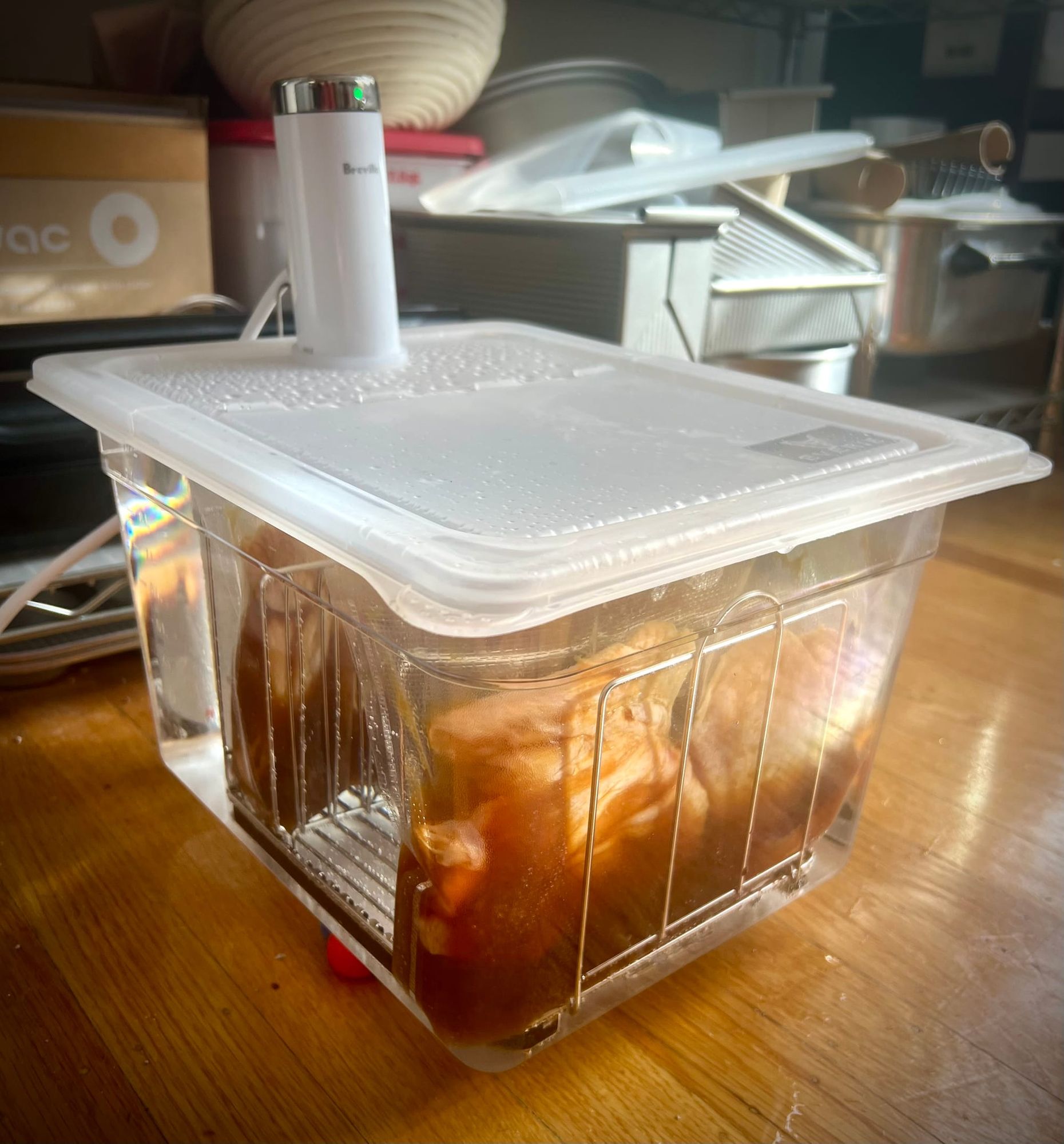 A clear plastic container is filled with water and bags of cooking chicken. A white Breville Joule sous vide appliance sticks up through the container's lid.