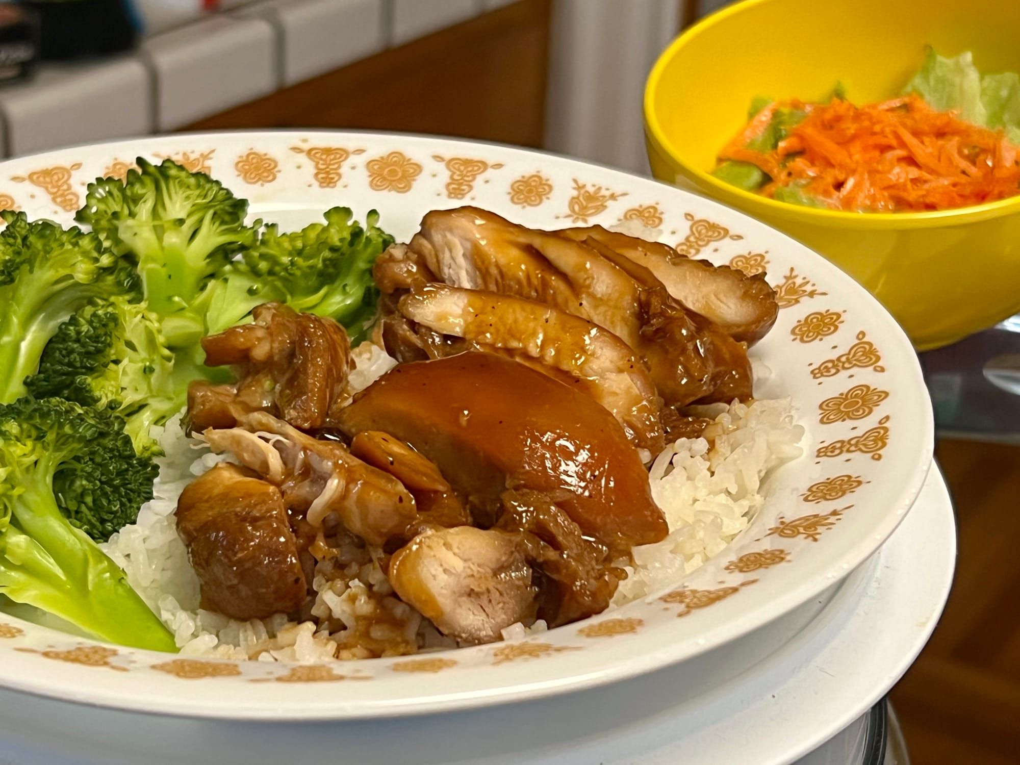 Sliced chicken is covered with a brown sauce and laid on a bed of rice, with broccoli on the side and a small iceberg & carrot salad.