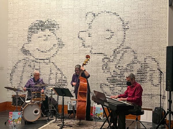 A jazz trio—drummer, upright bass, and piano—in front of a mural of Charlie Brown & Lucy, made of hundreds of Peanuts comics.