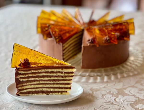 A slice of cake with seven thin layers and chocolate frosting, topped with a triangle of translucent shiny caramel. The whole cake is in the background.