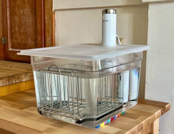 A clear plastic tub holds a Breville Joule immersion circulator. The tub has a cover, and inside there is a metal rack.