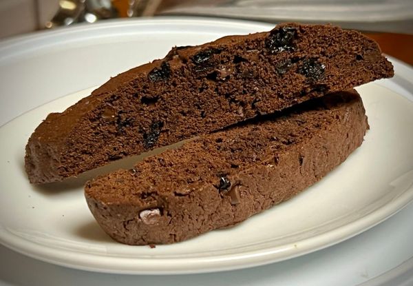 Two dark brown biscotti cookies are on a plate. They are studded with chopped dried cherries and mini chocolate chips.