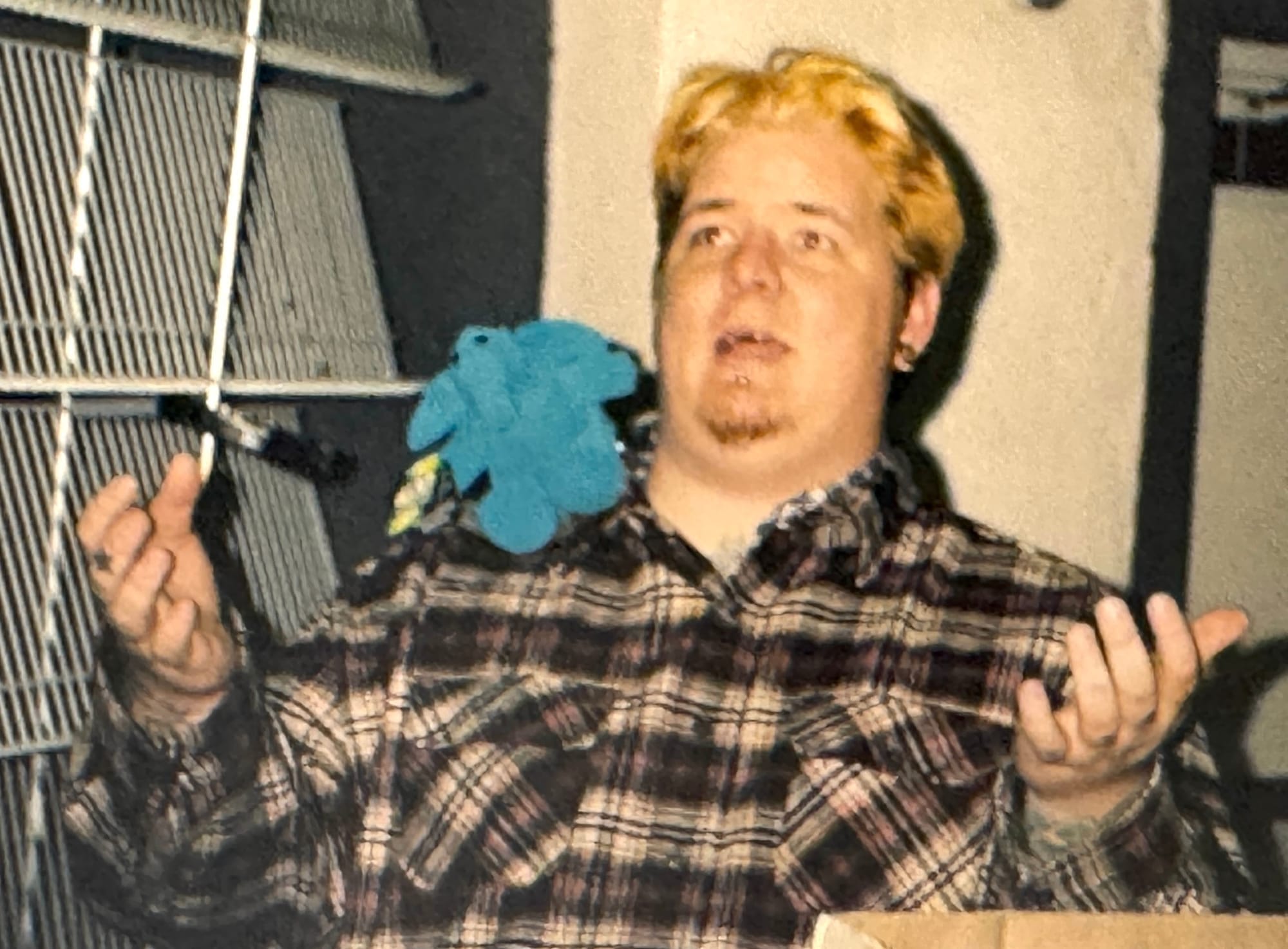 A man sitting with a silly look on his face and a plush toy on his shoulder. He has bleached ends and a flannel shirt and terribly thin hair on his chin.