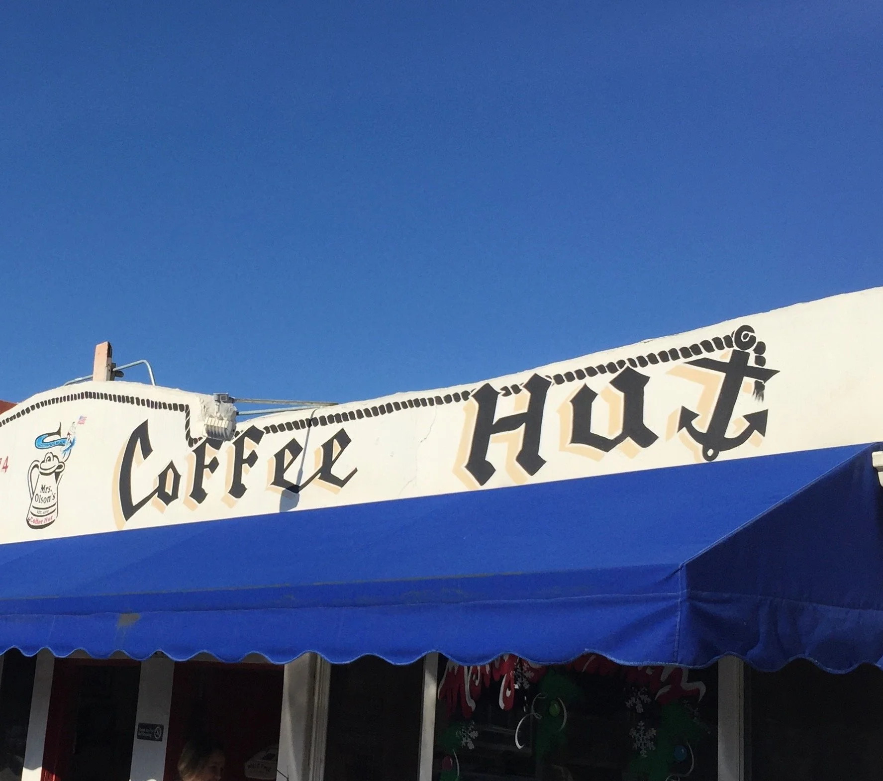 A partial store front for a coffee shop (restaurant, not drink stop) called "Coffee Hut". The sky is clear and blue, almost perfectly matching the awning below the painted signage.