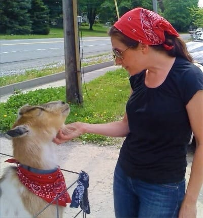 Alison petting a goat wearing a red bandana, both standing roadside on a sunny day