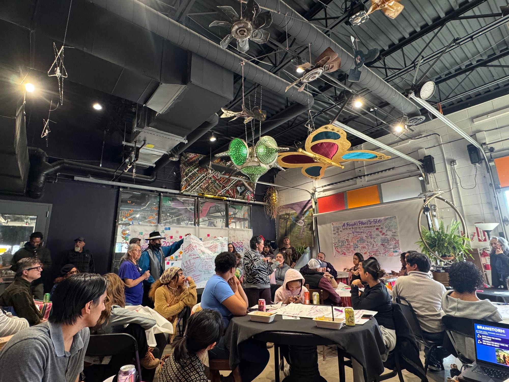 People are sitting around tables. A group of presenters holds a large sheet of paper with words and diagrams.