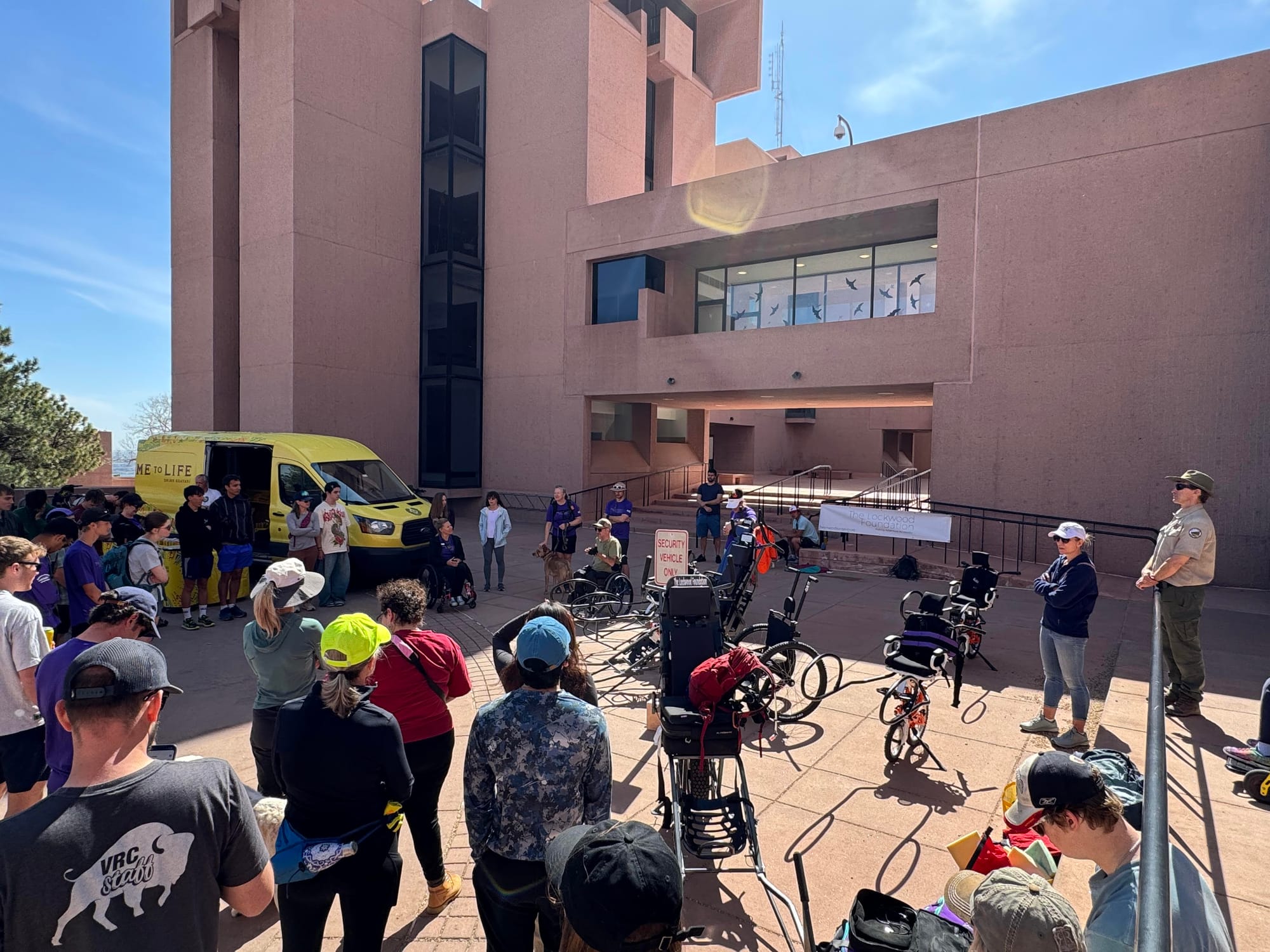 A group of people stand in front of the NCAR building, listening to people speaking.