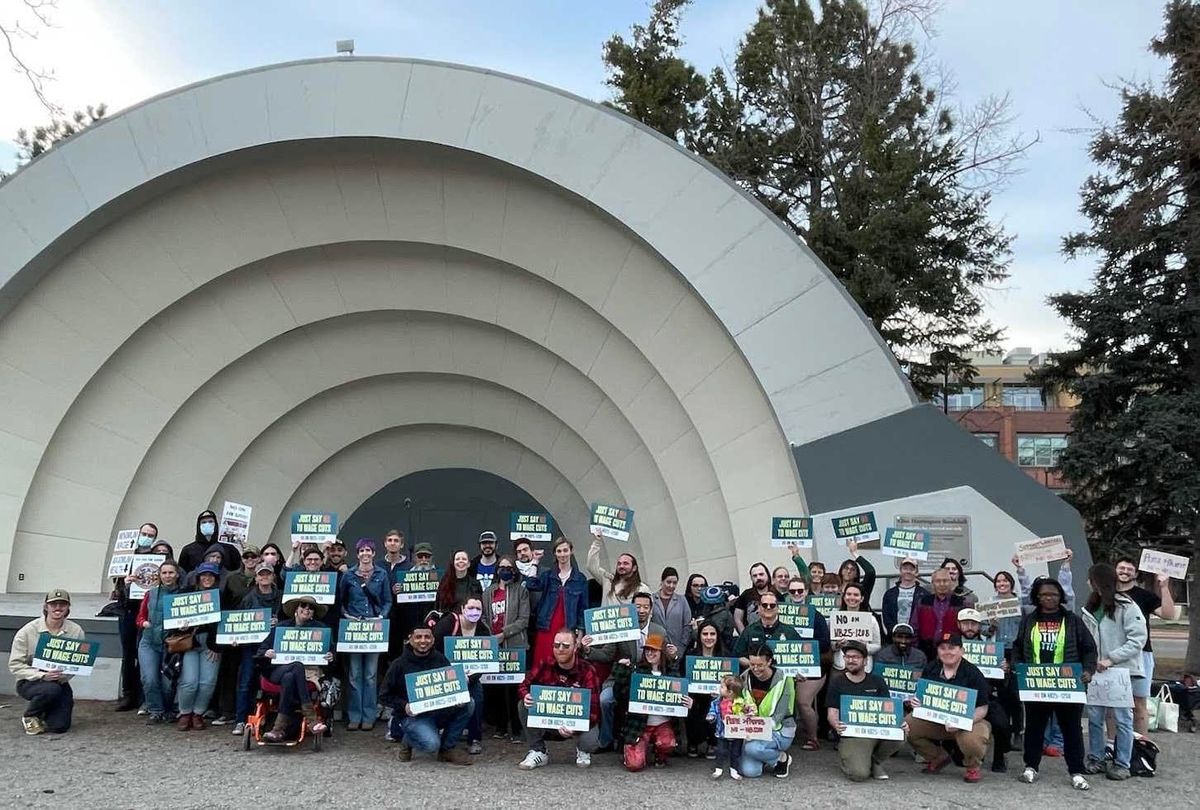 A large group of people holding "Just Say No to Wage Cuts" poses in front of the Boulder Bandshell.