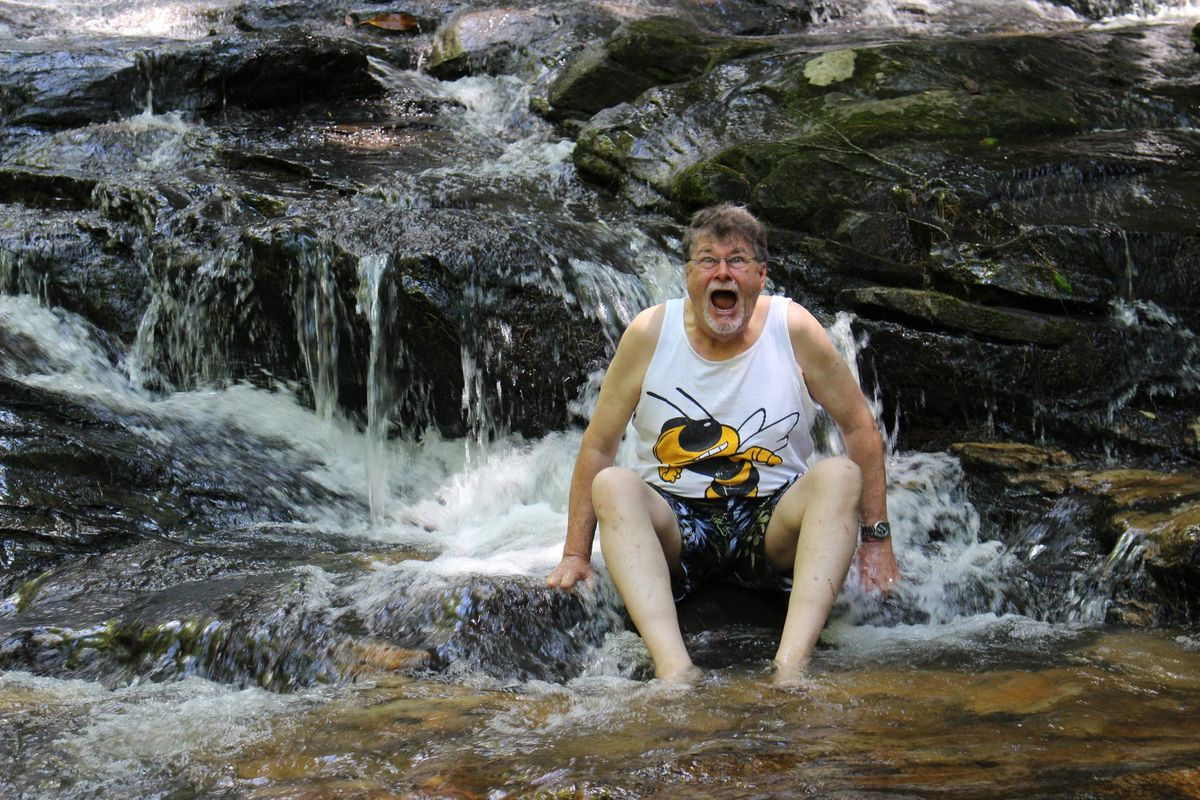An elderly man sits in a rushing creek, laughing with surprise as cold water splashes around him.