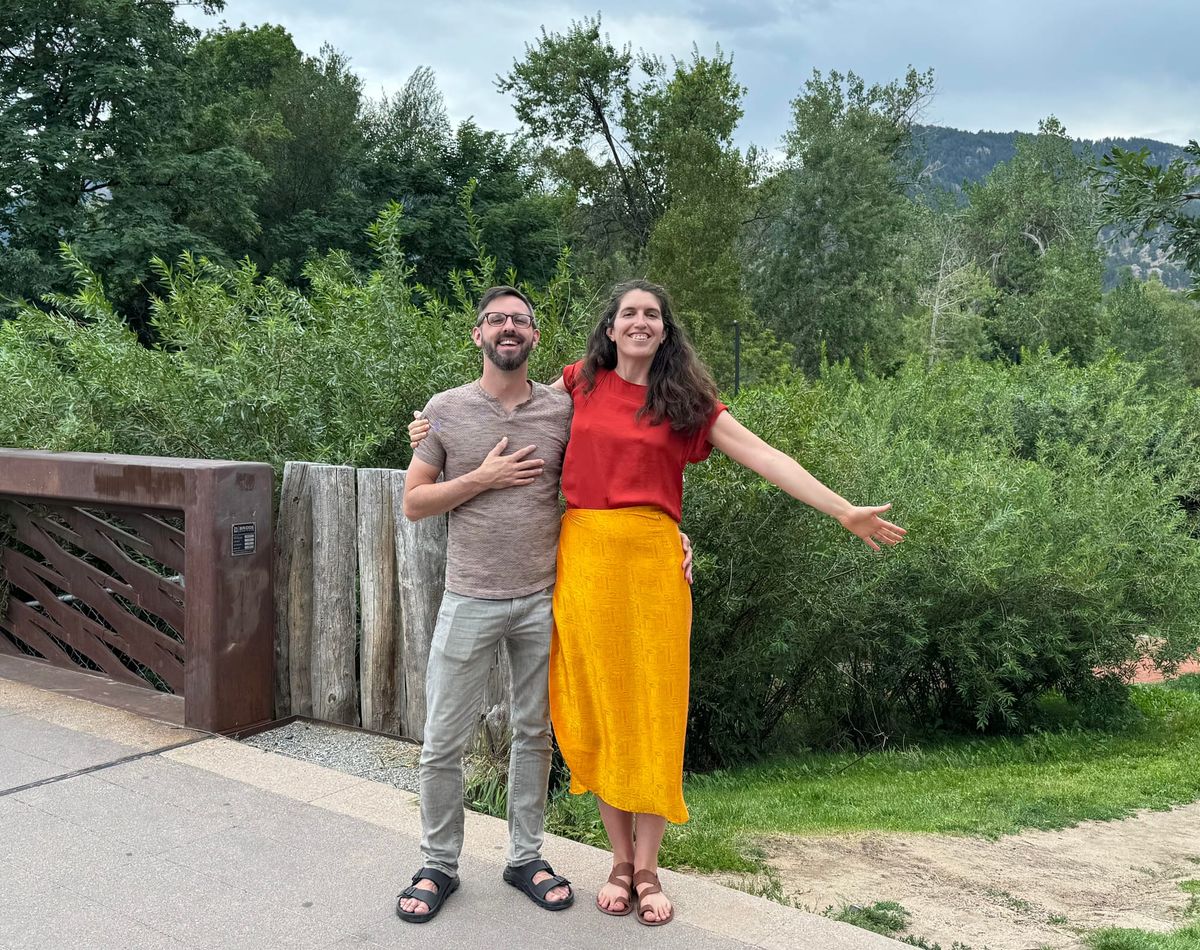 Two people pose near a pedestrian bridge downtown with metal railings and leafy trees in the background.