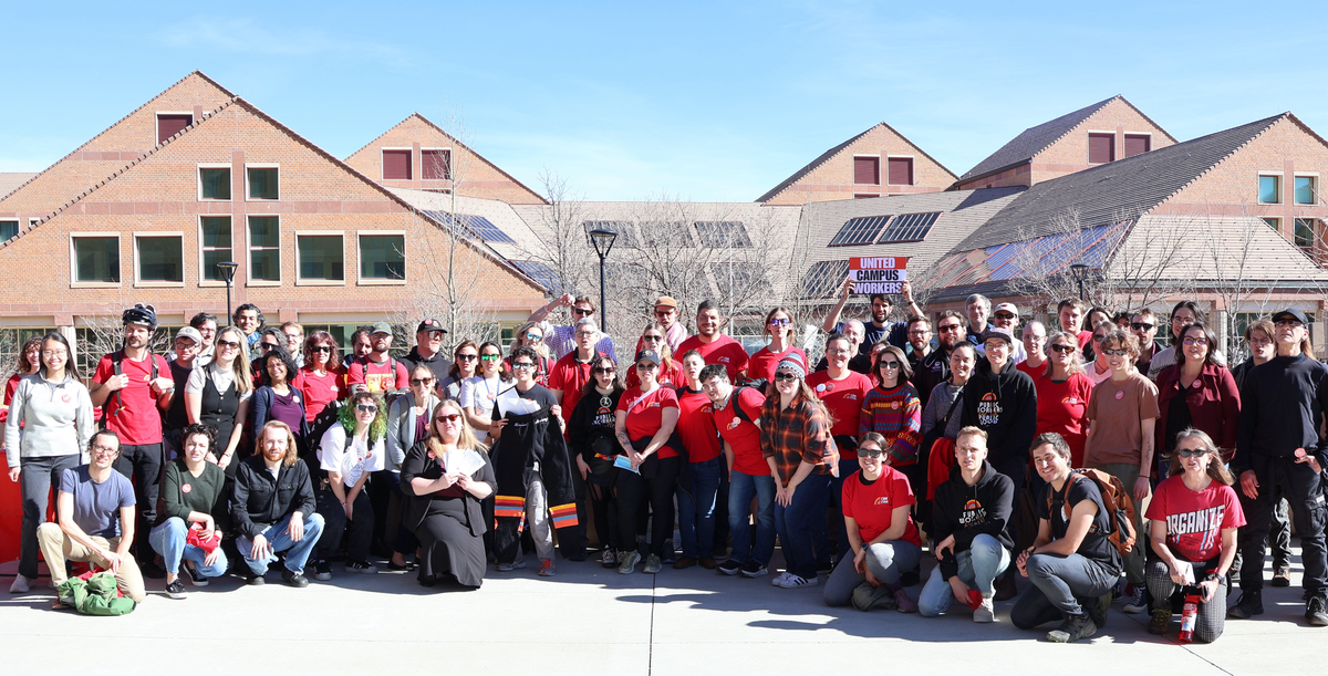 A large group of union members in red and black shirts stands in the sun outside the SEEC building.