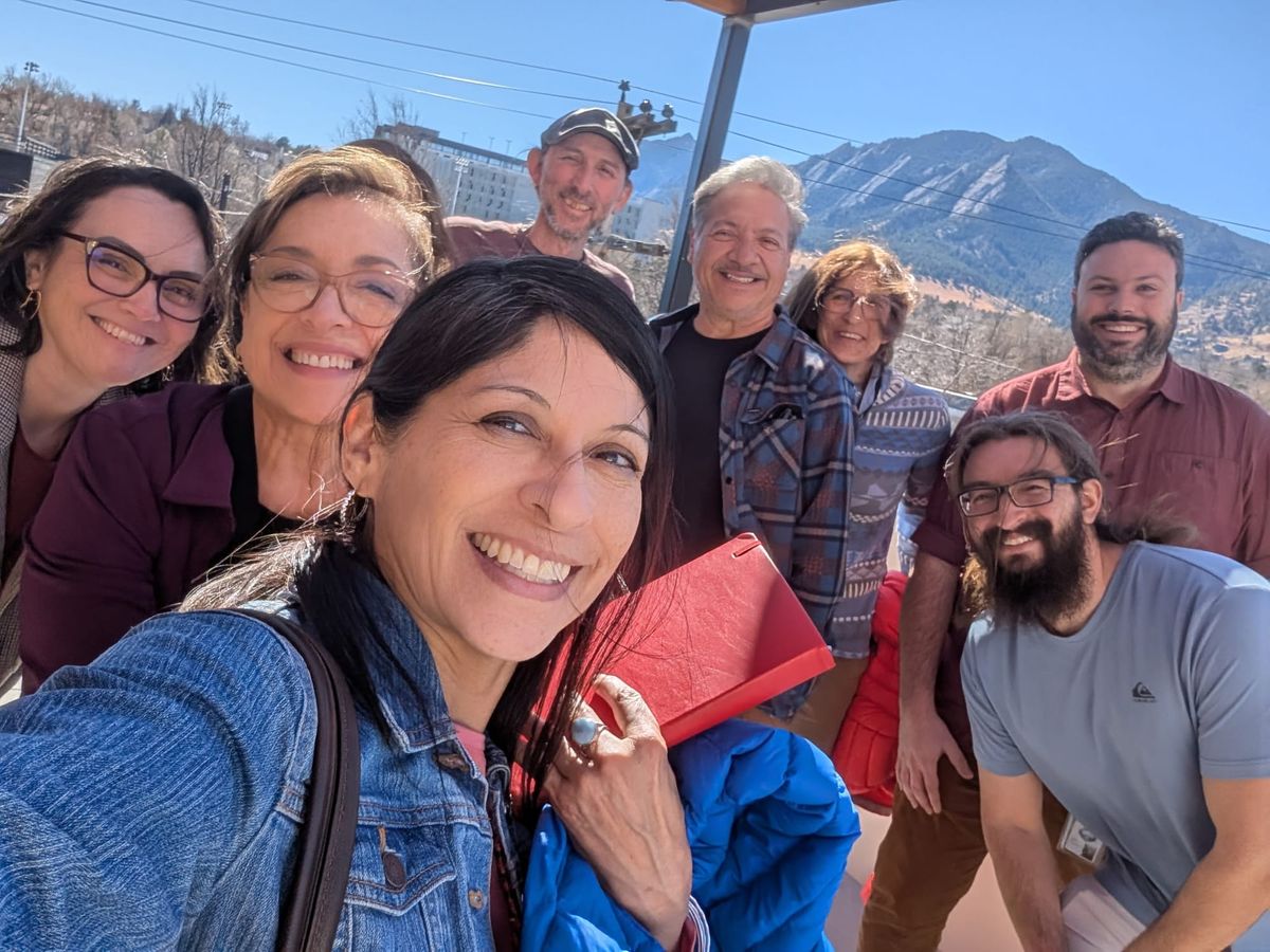 Eight people pose for a selfie on the rooftop deck of KGNU's new building, with the flatirons in the background.