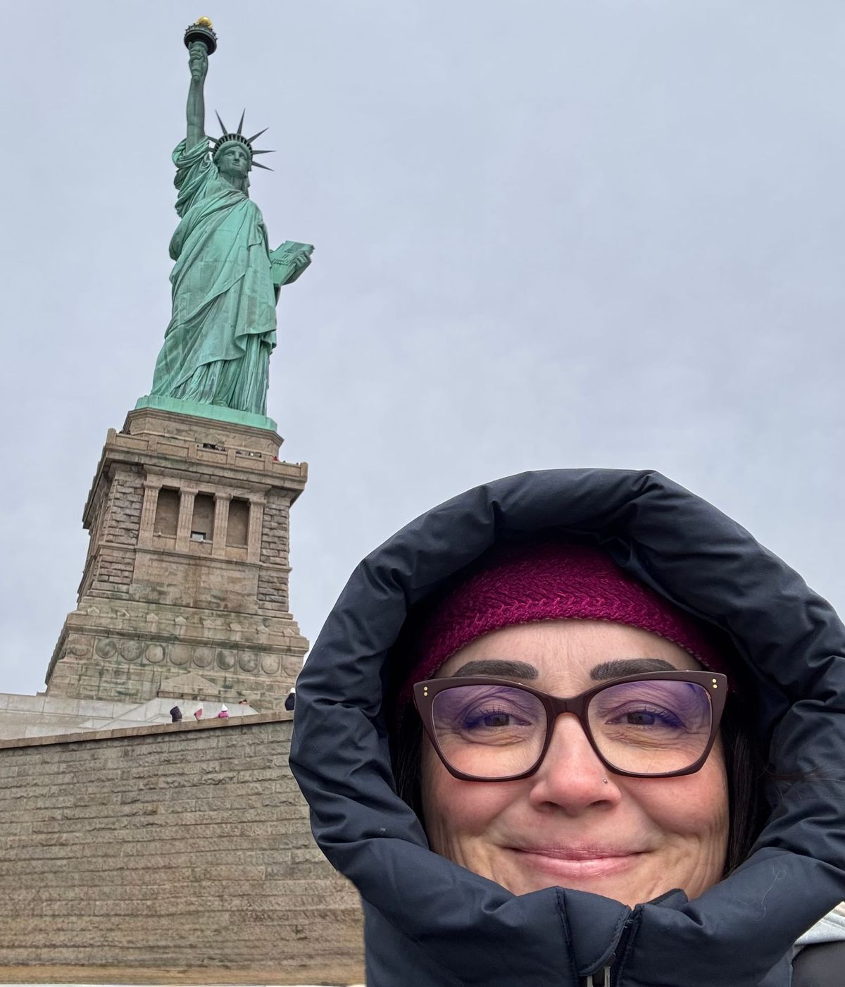 Nicole stands in front of the Statue of Liberty on an overcast day, wearing a winter hat and hooded jacket.