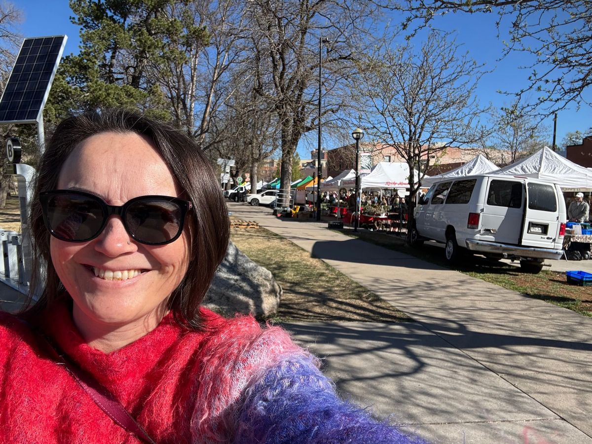 A person wearing sunglasses and a colorful sweater with vendor tents and a clear sky in the background.