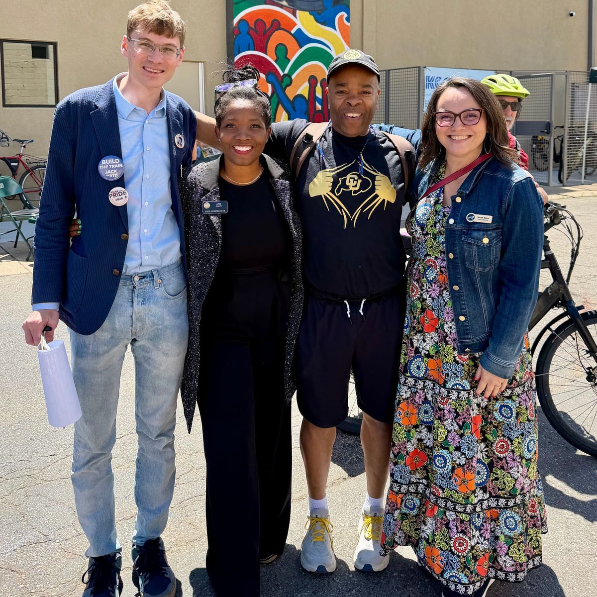 Four people stand together smiling in front of a colorful mural in the parking lot of KGNU's new downtown space. 
