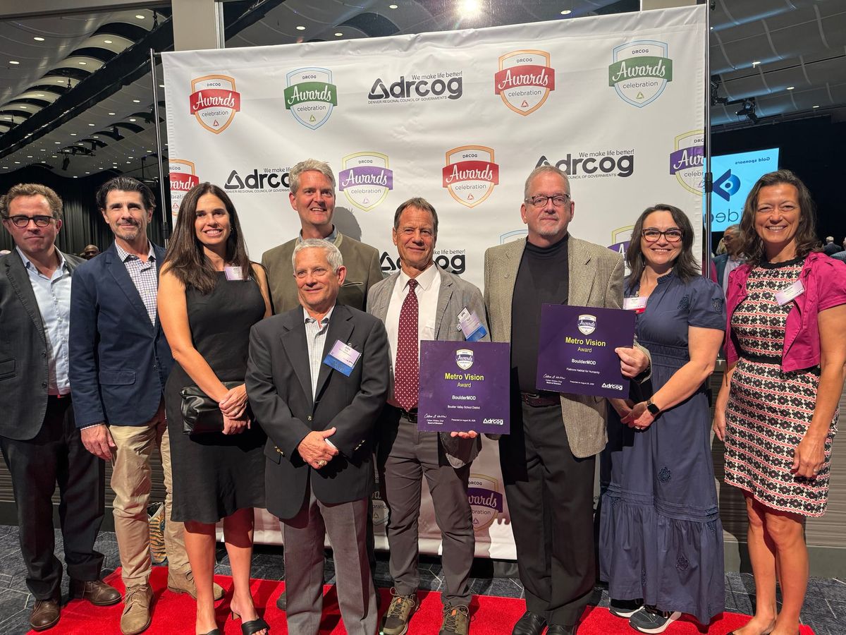 Nine people stand on a red carpet in front of a banner that reads "DRCOG Awards Celebration."