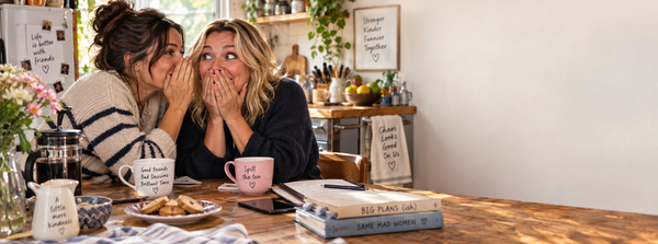 Two friends sharing a secret over coffee and biscuits at a kitchen table