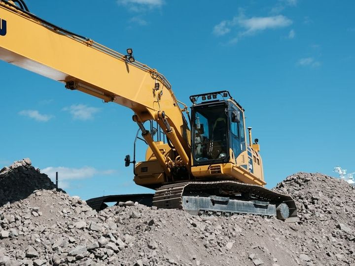An autonomous yellow excavator digs on a dynamic construction site, while a construction manager remotely monitors its progress and safety from a tablet computer.