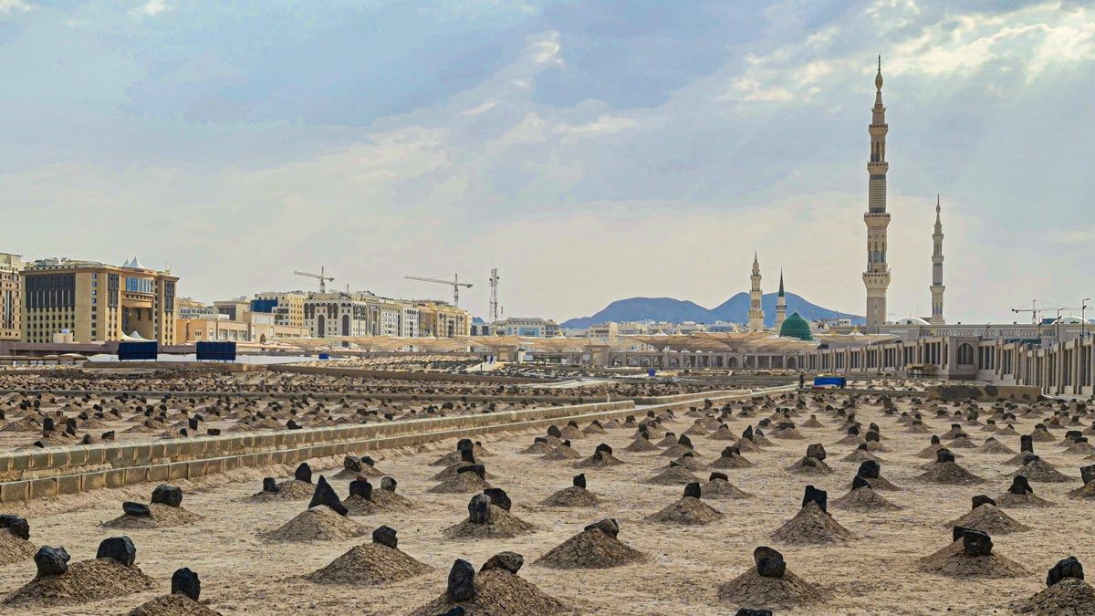 Rows of simple unmarked graves at Jannat al-Baqi cemetery in Medina under a bright cloudy sky, with the minarets of the Prophet's Mosque in the distance
