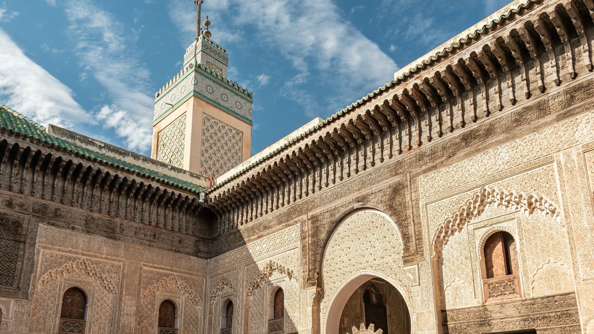 Carved cedar and stucco façade of the Bou Inania Madrasa courtyard in Fez, Morocco, with its green-tiled minaret against a bright sky