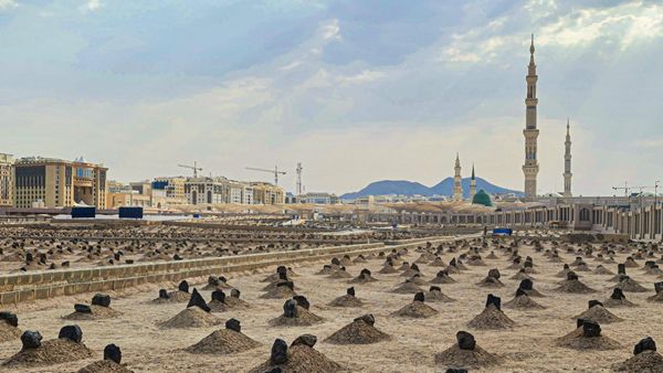Rows of simple unmarked graves at Jannat al-Baqi cemetery in Medina under a bright cloudy sky, with the minarets of the Prophet's Mosque in the distance