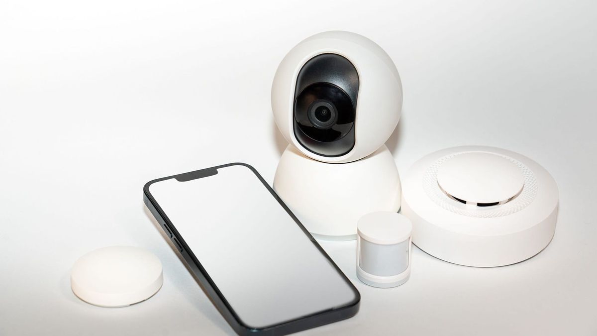 Smartphone beside a motion sensor, indoor camera, and other smart alarm sensors on a white surface