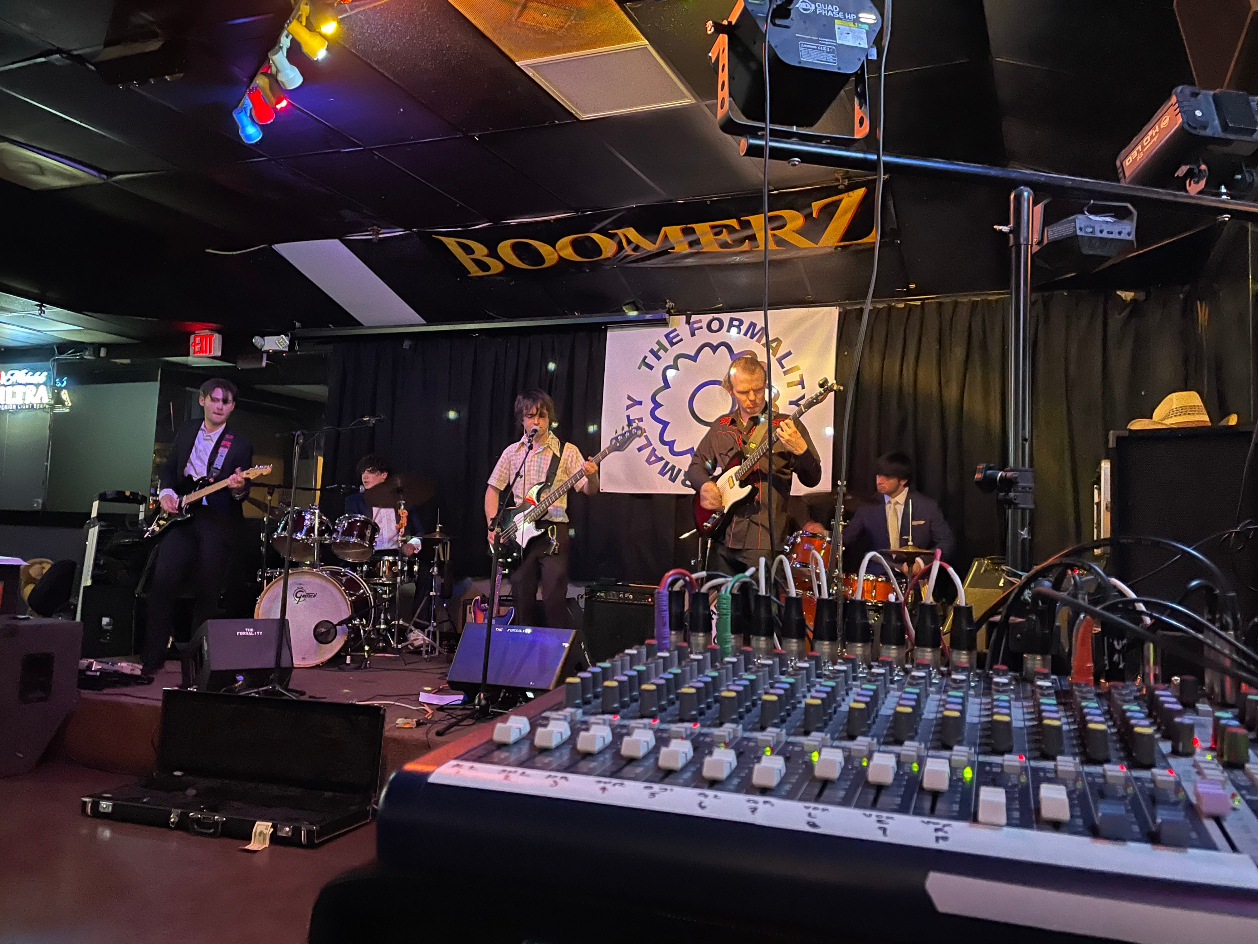 A band, left to right: guitarist, drummer, singing bassist, guitarist, drummer. On a stage in a bar called Boomerz. A banner of the band's name, The Formality, hangs on the wall behind the band. In the foreground, a mixing board. 
