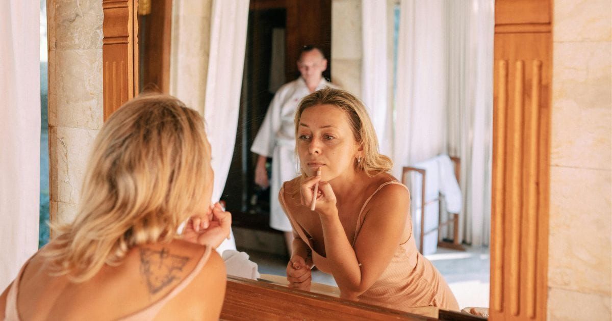 Woman applying makeup at mirror while husband watches in the background