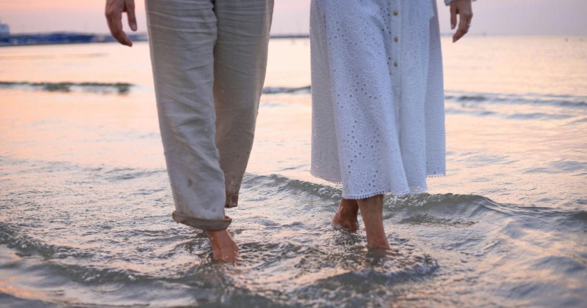 Couple standing barefoot together at ocean's edge at sunset