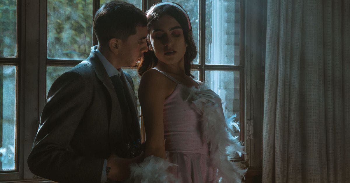 Man in suit leaning close to woman in pink feather dress by window suggesting dominance and surrender