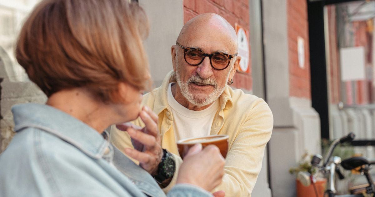 Older couple leaning in toward each other over coffee in conversation outside a cafe