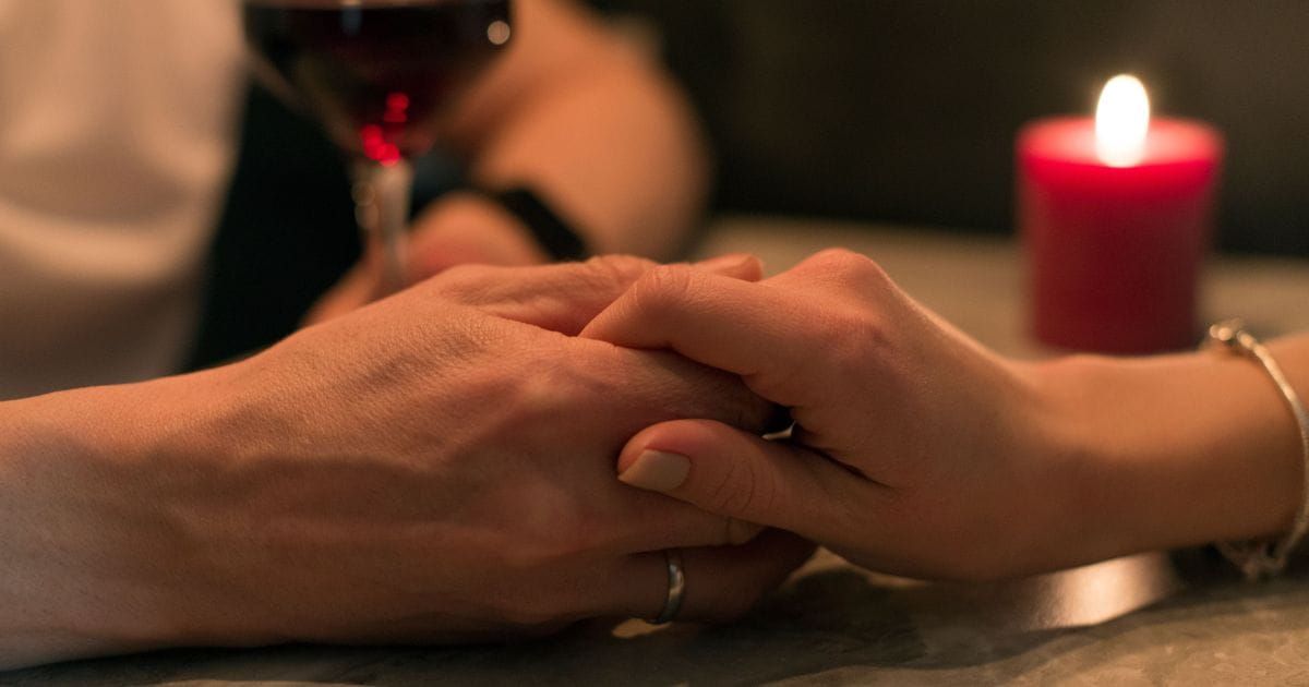 Two hands intertwined on a table with a red candle and wine glass in the background