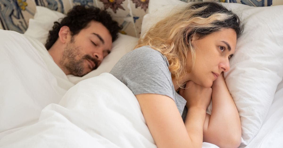 Woman lying awake in bed beside her sleeping partner, a quiet look of distance on her face