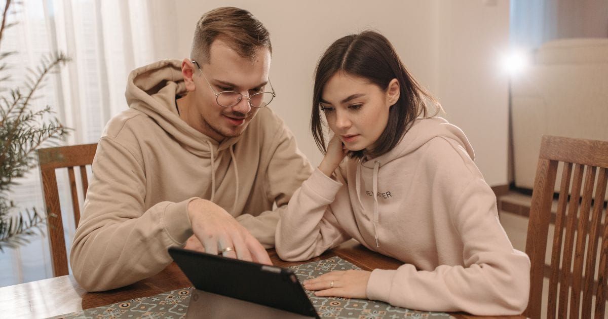 Couple looking closely at a tablet together at a table, wedding rings visible.