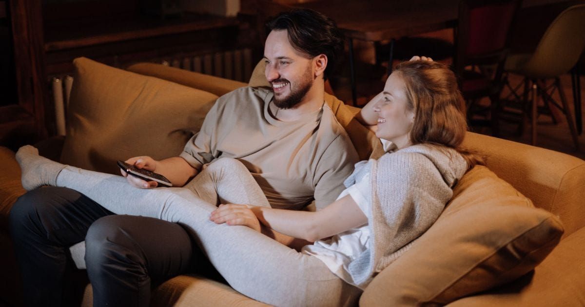 Couple on a couch smiling and looking at each other, hand resting on shoulder.