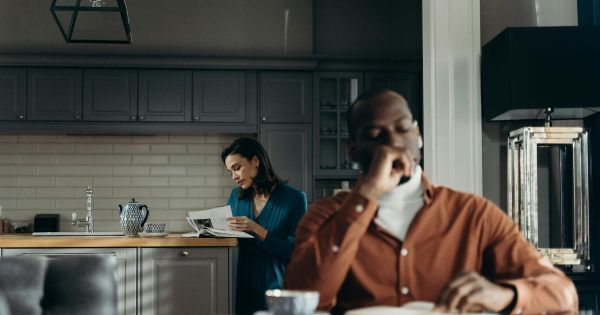Man sitting at kitchen table thoughtfully while woman reads in background