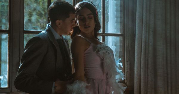  Man in suit leaning close to woman in pink feather dress by window suggesting dominance and surrender