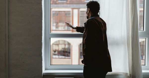Man standing at window looking out lost in thought with hand resting on glass