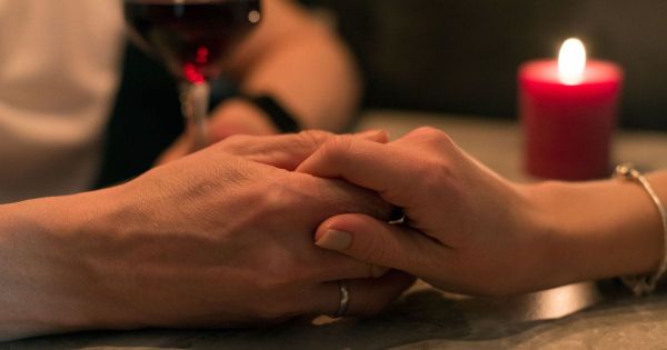 Two hands intertwined on a table with a red candle and wine glass in the background
