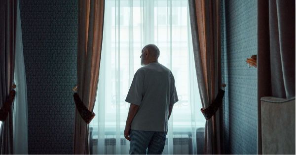 Older man standing at a curtained window seen from behind, looking out in quiet contemplation