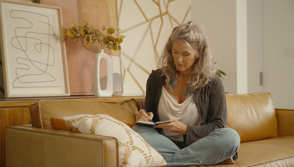 Older woman sitting cross-legged on a couch, writing in a journal